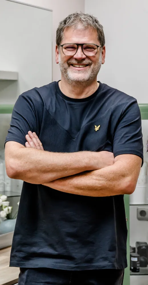 Dir Hermanns mit kurzen Haaren und Brille, der ein schwarzes T-Shirt trägt, lächelt und hat die Arme vor der Brust verschränkt. Er steht in einem Raum mit Regalen und Schränken im Hintergrund.
