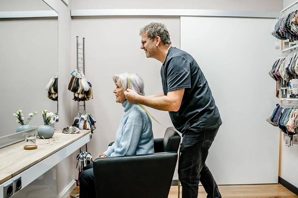 Friseur Dirk Hermanns hilft einer älteren Frau in einem Friseursalon beim Anprobieren einer Perücke. Die Frau sitzt in einem schwarzen Stuhl und trägt einen blauen Pullover. Der Mann steht lächelnd hinter ihr und hält ein Maßband in der Hand. An der Wand hängen verschiedene Perücken.